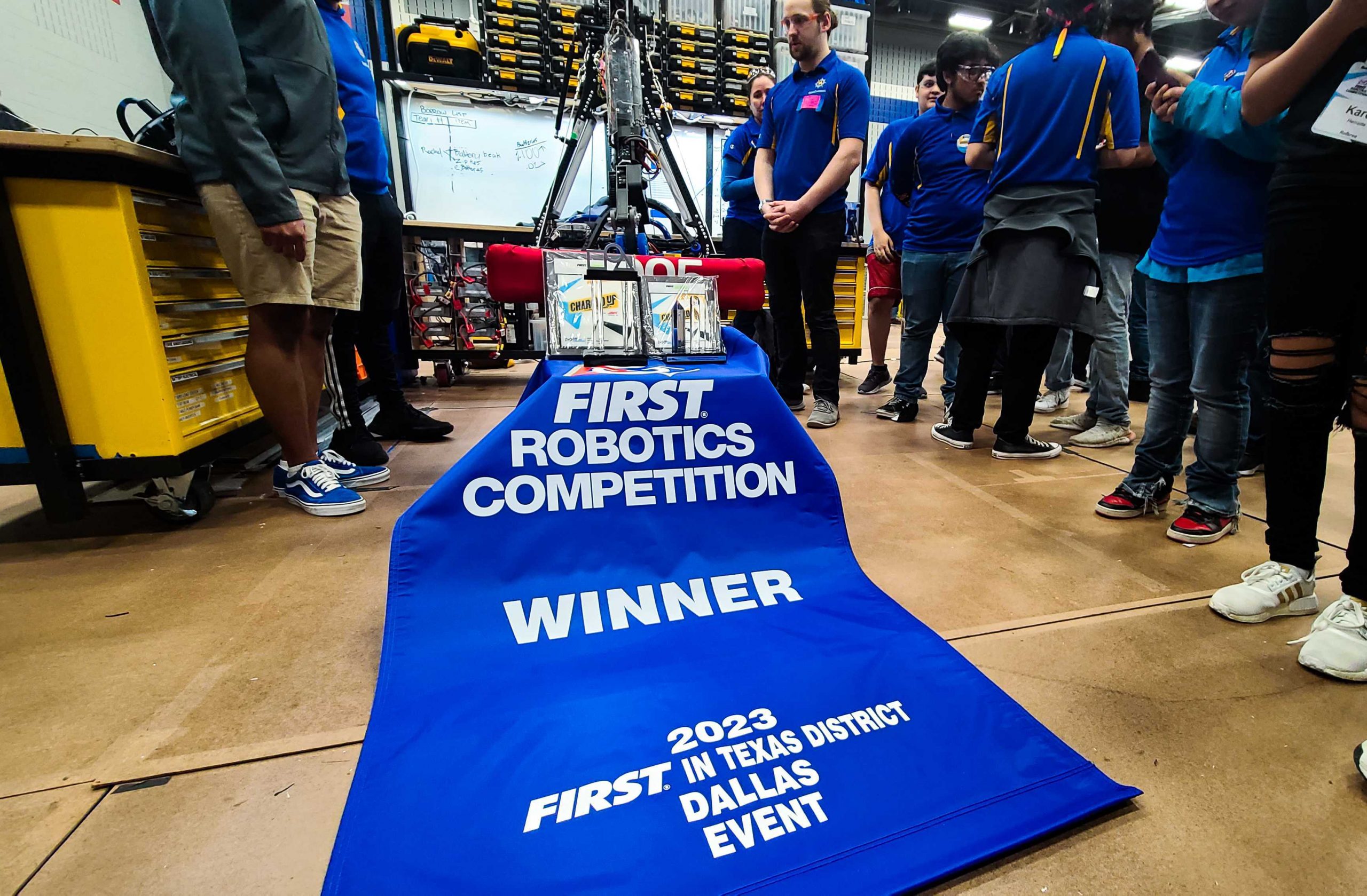 Image shows 3005's 2023 robot Amp sitting on its robot cart with a blue winners banner draped across the front. Two trophies and two plaques are sitting on the banner for winning the event and winning the district Excellence in Engineering award
